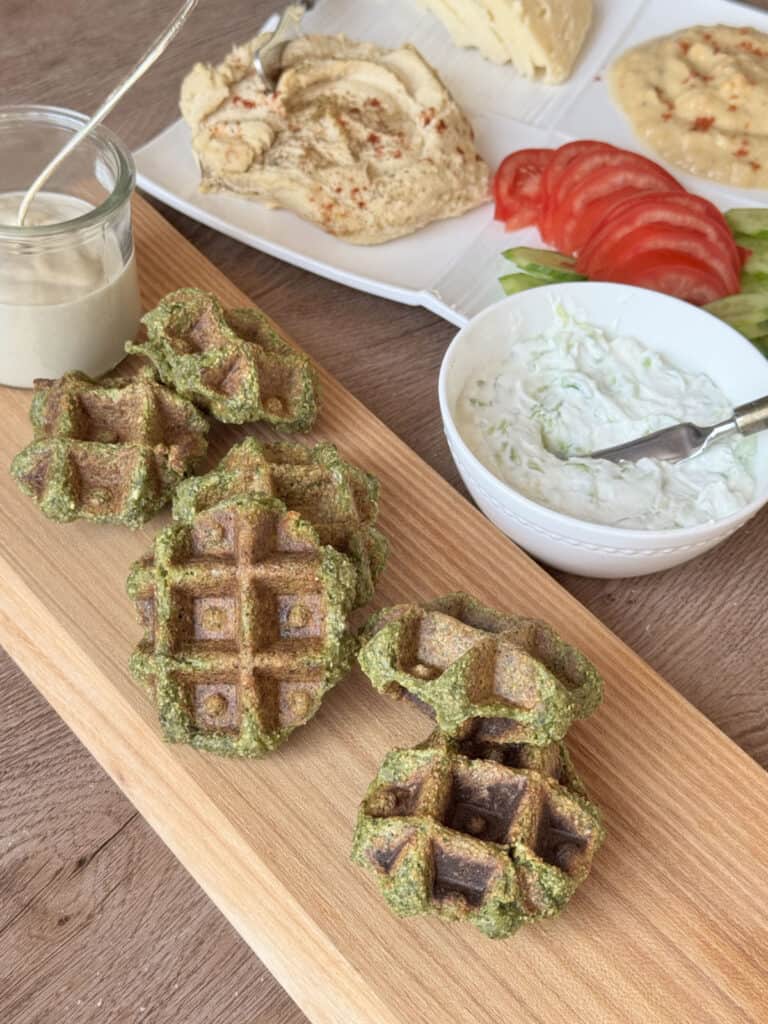 Falafel waffles on a board with mutabal, hummus, and tahini sauce. 
