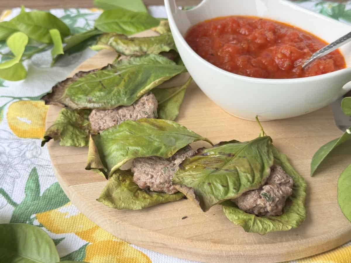 The meatballs in lemon leaves are arranged on a wooden board with red tomato sauce.