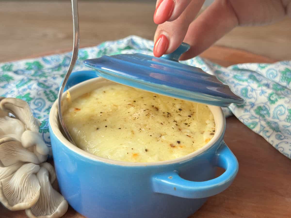 Creamy mushroom gratin served in a blue ceramic dish, with a metal spoon and fresh mushrooms nearby, on a wooden table.